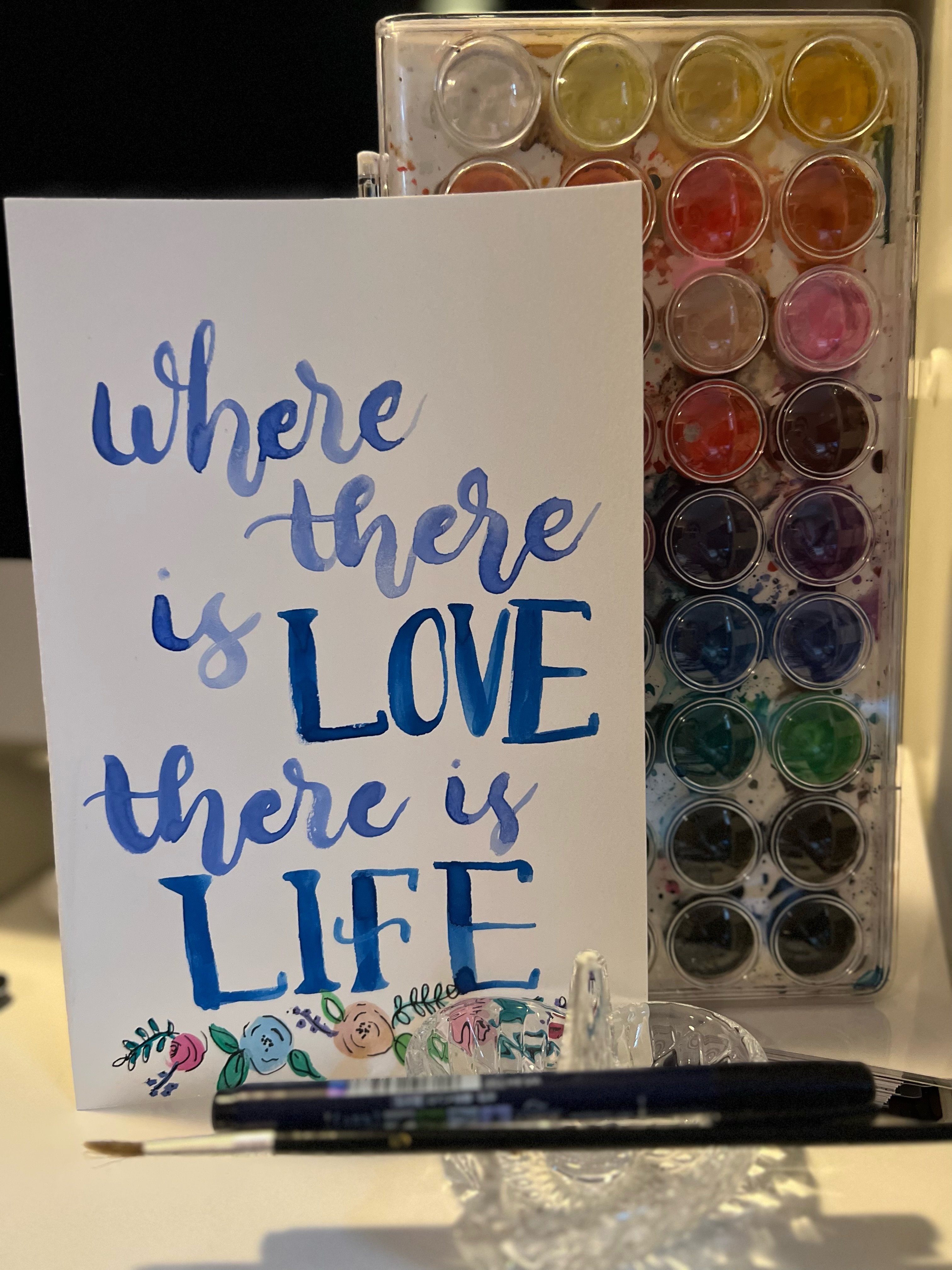 White card with blue watercolor writing that says "Where there is love there is life" with small watercolor painted flowers at bottom.