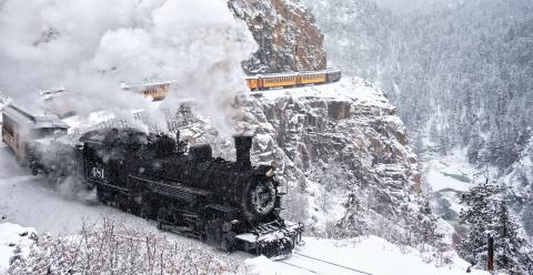 a black locomotive pulling yellow train cars through the snow on a mountainside